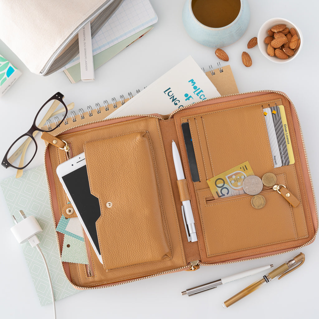  An opened pebbled leather travel wallet in camel on a white desk with pens, books, coffee and reading glasses.